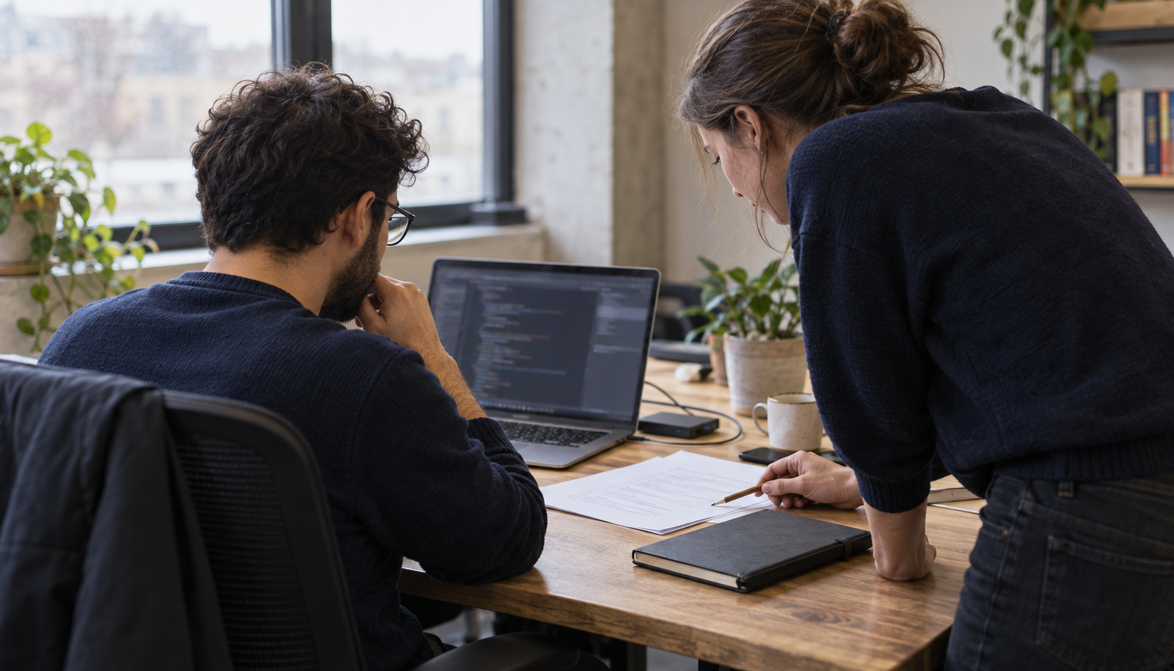 Two engineers review a software change and printed checklist beside a laptop in a naturally lit office.