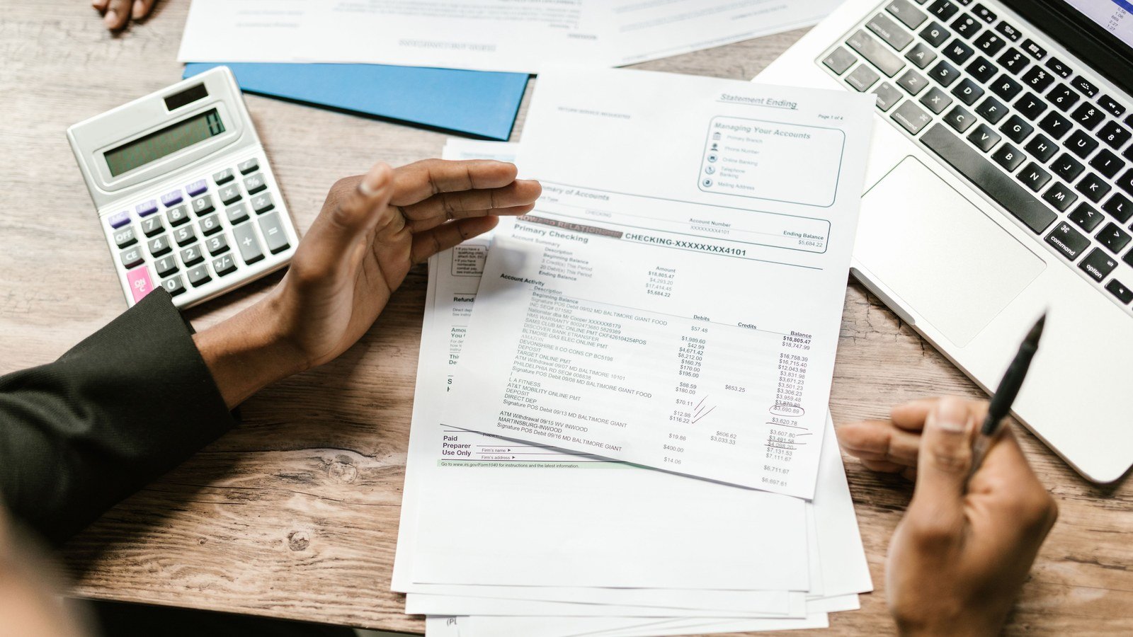 Hands holding printed financial documents beside a calculator and an open laptop on a wooden desk