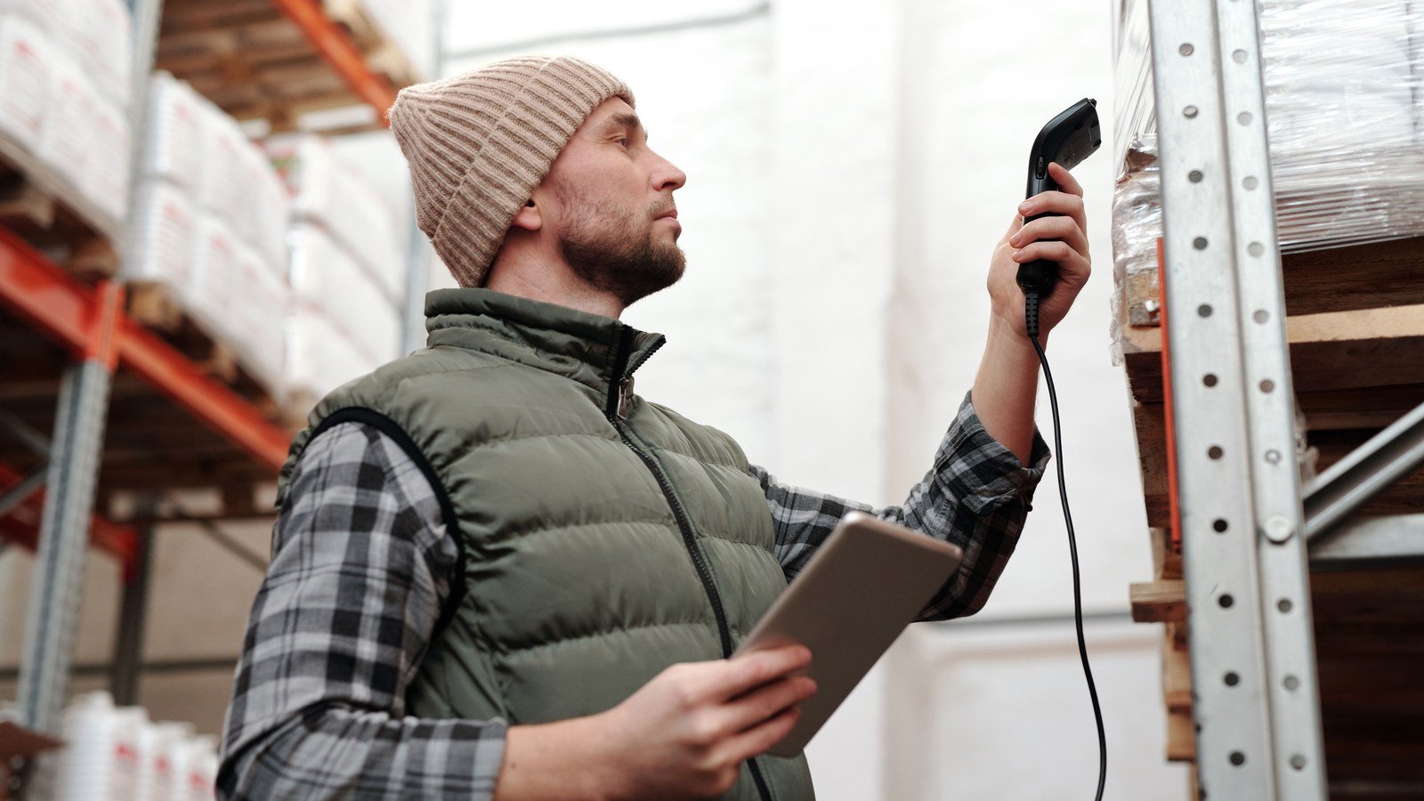 A warehouse worker in a knit cap and vest scans stock on a shelf with a handheld barcode scanner while holding a tablet