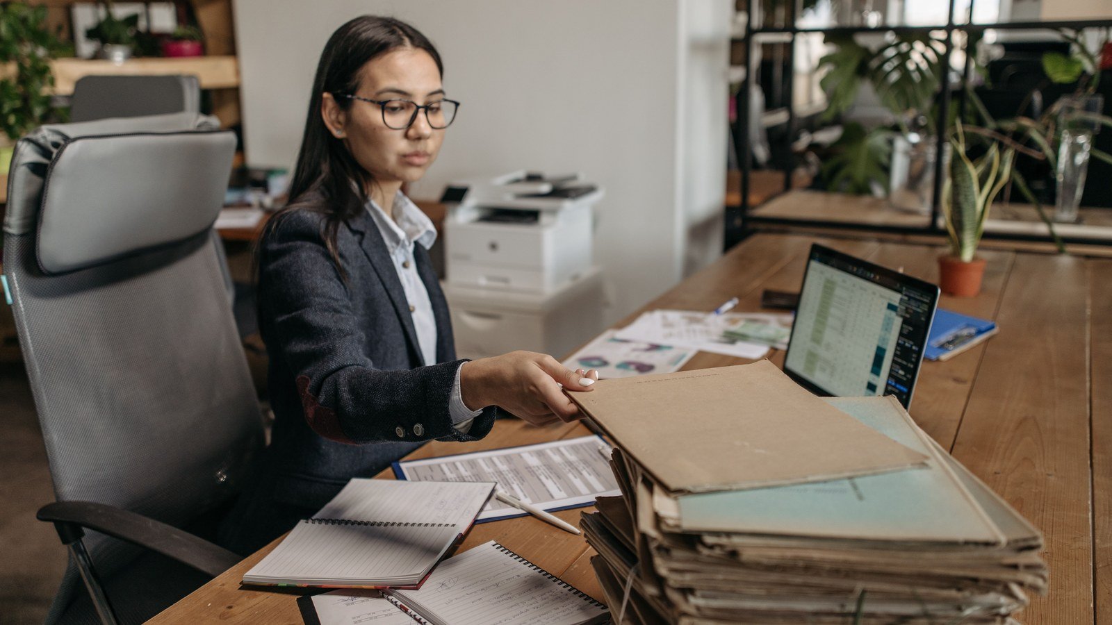 A woman in an office works on a laptop beside a tall stack of paper folders and loose documents on a wooden desk